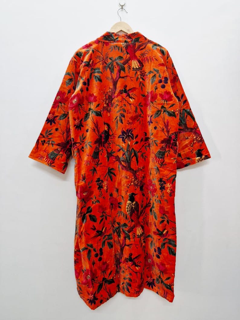 May include: An orange velvet robe with a vibrant floral and bird pattern. The long-sleeved robe features a detailed design of flowers, leaves, and birds in shades of red, green, and black. The robe is hanging on a wooden hanger.