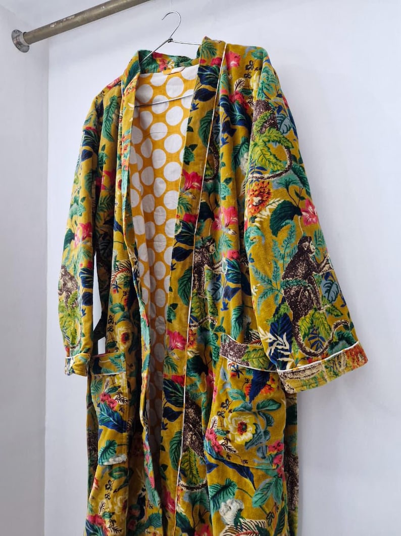 May include: A long, mustard-yellow robe with a vibrant floral and monkey print. The robe features a white polka dot lining and white trim along the edges. It is hanging on a metal rod against a white wall.