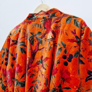 May include: An orange velvet robe with a floral and bird design. The robe has a collared neckline and long sleeves. The pattern includes red flowers, green leaves, and colourful birds, creating a detailed appearance.