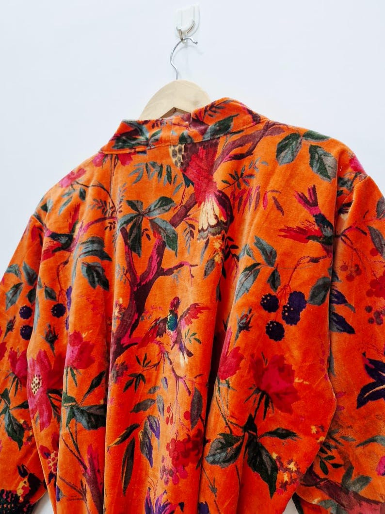 May include: An orange velvet robe with a floral and bird design. The robe has a collared neckline and long sleeves. The pattern includes red flowers, green leaves, and colourful birds, creating a detailed appearance.