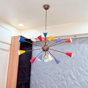 Puede incluir: Una lámpara de araña moderna de mediados de siglo con acabado bronce y múltiples pantallas cónicas de colores. Las pantallas son de varios colores, incluyendo rojo, amarillo, azul y morado. La lámpara está suspendida del techo.