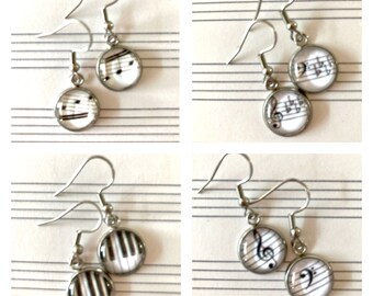Peut inclure: Quatre paires de boucles d'oreilles pendantes argentées avec des breloques rondes en verre représentant des notes de musique et des touches de piano. Les breloques sont blanches avec des notes de musique et des touches de piano noires.