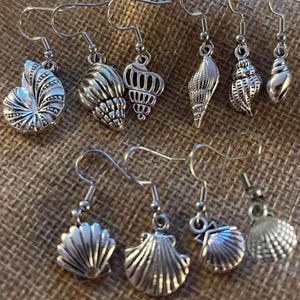 May include: A collection of ten silver-toned metal earrings, each featuring a unique seashell or conch design. The earrings are equipped with standard hook-style ear wires for hanging. Each charm displays intricate textures and details of various sea shells.