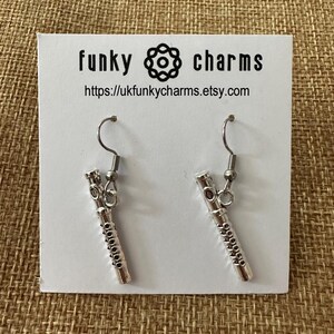 Peut inclure: Boucles d'oreilles pendantes argent&eacute;es avec un motif de fl&ucirc;te miniature. Les boucles d'oreilles sont sur une carte blanche avec le texte "funky charms" et une adresse de site web.