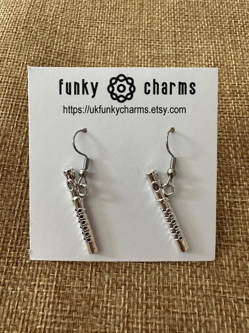 Peut inclure: Boucles d'oreilles pendantes argent&eacute;es avec un motif de fl&ucirc;te miniature. Les boucles d'oreilles sont sur une carte blanche avec le texte "funky charms" et une adresse de site web.