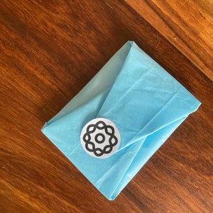 Peut inclure: Une enveloppe en papier de soie bleu clair avec un autocollant circulaire noir et blanc repr&eacute;sentant un motif stylis&eacute;.