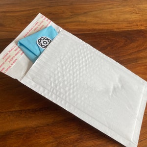 Peut inclure: Une enveloppe matelass&eacute;e blanche avec un tissu bleu pli&eacute; &agrave; l'int&eacute;rieur. L'enveloppe porte une &eacute;tiquette avec le texte "EXPOSE ADHESIVE" et "SECURE SEAL" r&eacute;p&eacute;t&eacute; plusieurs fois.
