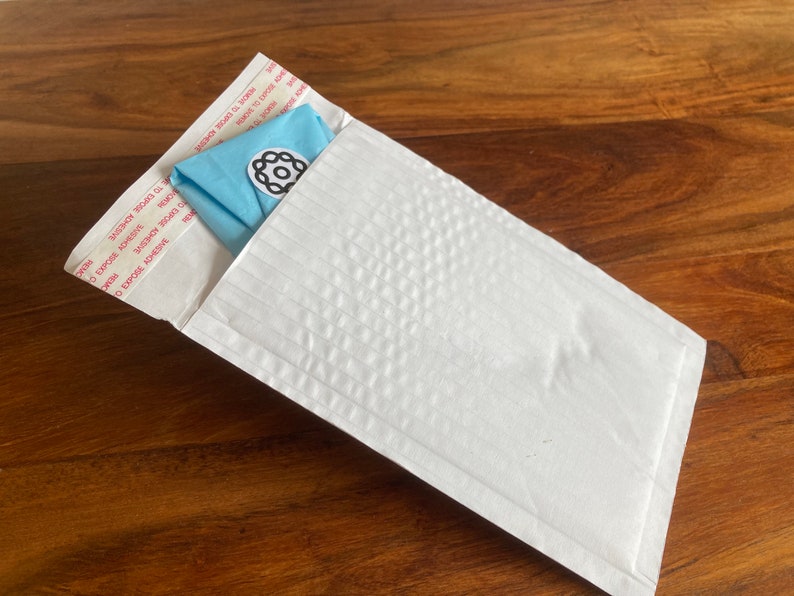 Peut inclure: Une enveloppe matelass&eacute;e blanche avec un tissu bleu pli&eacute; &agrave; l'int&eacute;rieur. L'enveloppe porte une &eacute;tiquette avec le texte "EXPOSE ADHESIVE" et "SECURE SEAL" r&eacute;p&eacute;t&eacute; plusieurs fois.