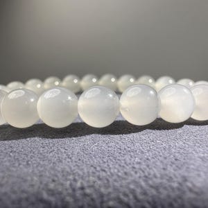 K&ouml;nnte beinhalten: Ein Armband bestehend aus einer einzelnen Reihe glatter, runder, halb-durchscheinender wei&szlig;er Perlen. Jede Perle hat eine polierte, gl&auml;nzende Oberfl&auml;che, die das Licht reflektiert und ein einheitliches Erscheinungsbild des gesamten Schmuckst&uuml;cks erzeugt.