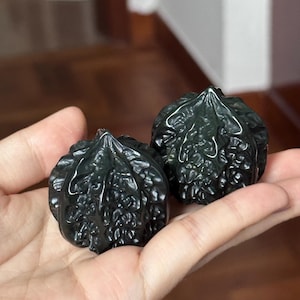 Pode incluir: Dois objetos decorativos em forma de noz, verde escuro. Os objetos têm uma superfície texturizada com detalhes intrincados e um acabamento brilhante. Os objetos são segurados na mão de uma pessoa, com um fundo desfocado.