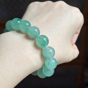 Pode incluir: Uma pulseira composta por contas lisas, redondas e translúcidas de cor verde-clara. As contas exibem tons variados de verde com padrões internos subtis semelhantes a nuvens e uma superfície polida e refletora, unidas num círculo contínuo.