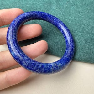 Könnte beinhalten: Ein lebendiges blaues Armreif-Armband aus Edelstein. Das Armband ist ein massives, kreisförmiges Band mit polierter, glänzender Oberfläche. Die Farbe ist ein tiefes, sattes Blau mit helleren, gesprenkelten Mustern. Ein klassisches Accessoire.