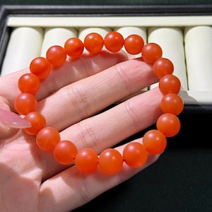 May include: A stretch bracelet composed of numerous smooth, round, vibrant orange beads. The beads have a matte, semi-translucent finish and are strung together on a flexible cord, forming a circular shape that fits around the wrist.