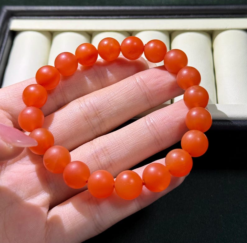 May include: A stretch bracelet composed of numerous smooth, round, vibrant orange beads. The beads have a matte, semi-translucent finish and are strung together on a flexible cord, forming a circular shape that fits around the wrist.