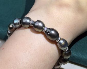 Op de afbeelding: Een donkergrijze parelarmband met diamantaccenten. De ovaalvormige parels zijn geregen met kleine, met diamanten bezette afstandhouders. De armband wordt om een pols gedragen.