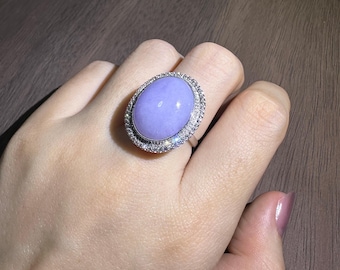 May include: A silver ring featuring a large, oval, lavender-colored gemstone. The gemstone is surrounded by a halo of small, sparkling clear stones. The ring is set on a silver band.