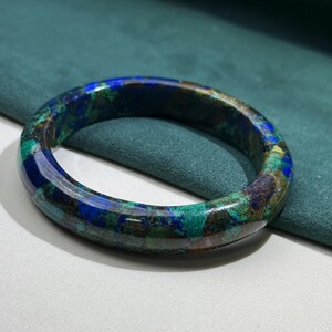 May include: A round, polished bracelet with a mosaic pattern of blue, green, and brown hues. The gemstone bracelet is set against a teal background, showcasing its natural colors and textures. This piece of jewelry is a unique accessory.
