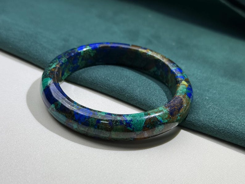 May include: A round, polished bracelet with a mosaic pattern of blue, green, and brown hues. The gemstone bracelet is set against a teal background, showcasing its natural colors and textures. This piece of jewelry is a unique accessory.