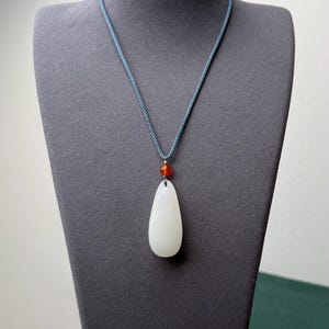 May include: A white teardrop-shaped pendant necklace with a small orange bead and a blue cord. The pendant is smooth and polished, and the cord is a woven style. The necklace is displayed on a gray mannequin.