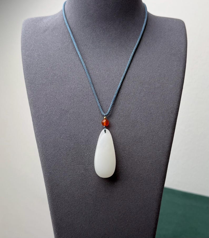 May include: A white teardrop-shaped pendant necklace with a small orange bead and a blue cord. The pendant is smooth and polished, and the cord is a woven style. The necklace is displayed on a gray mannequin.