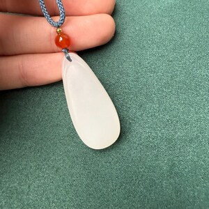 May include: A white teardrop-shaped pendant necklace with a blue cord. The pendant is smooth and opaque, with a small orange bead and a gold-colored bead above it. The background is a solid teal green.