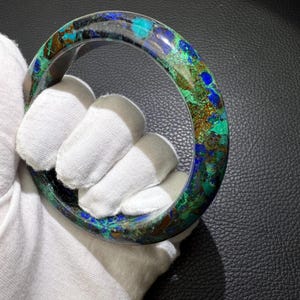 May include: A round, polished bracelet with a mosaic of colors including blue, green, and brown. The bracelet is held by a gloved hand, showcasing its smooth, curved surface. The gemstone bracelet is a unique piece of jewelry.