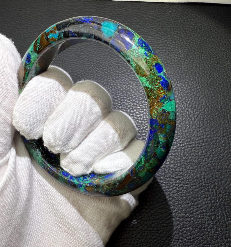 May include: A round, polished bracelet with a mosaic of colors including blue, green, and brown. The bracelet is held by a gloved hand, showcasing its smooth, curved surface. The gemstone bracelet is a unique piece of jewelry.