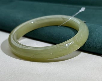 May include: A pale green jade bangle bracelet with carved floral designs. The smooth, polished surface reflects light, highlighting the intricate details of the carvings. The bracelet is displayed on a white and green background.