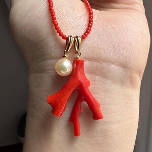Op de afbeelding: Een rode kralenketting met een koraal hanger en een parel. De koraal hanger heeft een heldere rode kleur en een takachtige vorm. De parel is cr&egrave;mewit en hangt aan een goudkleurige sluiting.