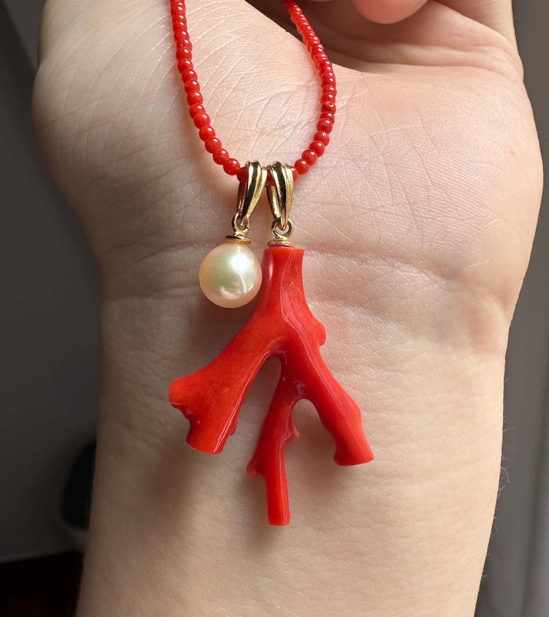 Op de afbeelding: Een rode kralenketting met een koraal hanger en een parel. De koraal hanger heeft een heldere rode kleur en een takachtige vorm. De parel is cr&egrave;mewit en hangt aan een goudkleurige sluiting.