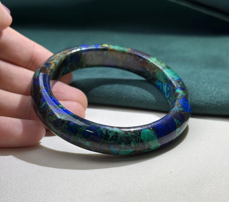 May include: A polished, round bracelet with a marbled appearance. The bracelet features a blend of deep blue, green, and brown hues. The gemstone bracelet is a unique piece of jewelry.