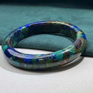 May include: A polished, circular bracelet with a marbled appearance. The bracelet features a blend of blue, green, and brown hues. The gemstone bracelet is displayed on a white surface with a teal background.