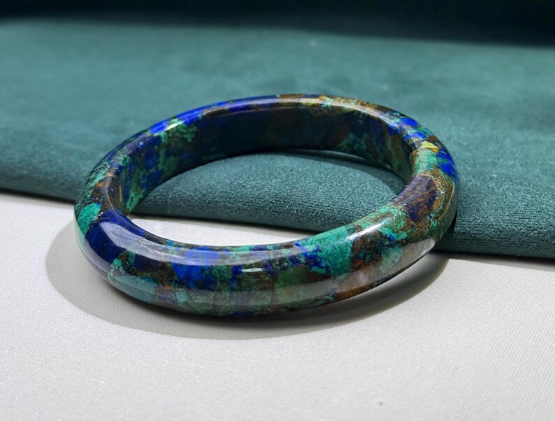 May include: A polished, circular bracelet with a marbled appearance. The bracelet features a blend of blue, green, and brown hues. The gemstone bracelet is displayed on a white surface with a teal background.