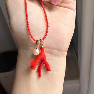 Op de afbeelding: Een rode kralenketting met een gouden hanger met een koraaltak en een parel. De koraaltak is felrood en de parel is cr&egrave;mewit. De ketting wordt gepresenteerd tegen een huidskleurige achtergrond.