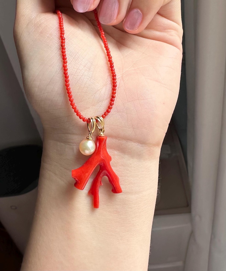 Op de afbeelding: Een rode kralenketting met een gouden hanger met een koraaltak en een parel. De koraaltak is felrood en de parel is cr&egrave;mewit. De ketting wordt gepresenteerd tegen een huidskleurige achtergrond.