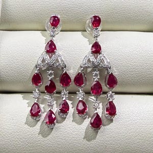 May include: A pair of elegant chandelier earrings featuring vibrant red pear-shaped gemstones and sparkling white accent stones. The earrings have a silver-colored metal setting and a cascading design, perfect for special occasions.