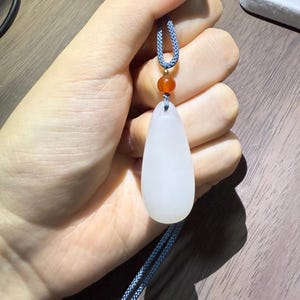 May include: A white teardrop-shaped pendant on a blue cord, accented with a small orange bead. The pendant is smooth and opaque, held against a neutral background. The cord is a twisted, braided design.