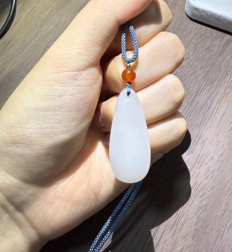 May include: A white teardrop-shaped pendant on a blue cord, accented with a small orange bead. The pendant is smooth and opaque, held against a neutral background. The cord is a twisted, braided design.