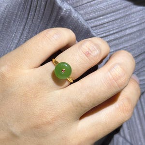 May include: A gold ring featuring a circular, smooth green jade stone centerpiece with a small, round, reddish-pink gemstone set in the middle. The gold band has a textured, leaf-like pattern on both sides of the central jade disc setting.