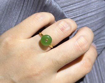 Könnte beinhalten: Ein Goldring mit einem runden, glatten grünen Jadestein als Mittelstück, in dessen Mitte ein kleiner, runder, rötlich-rosa Edelstein gefasst ist. Die Goldschiene weist auf beiden Seiten des zentralen Jade-Elements ein blattartiges Muster auf.