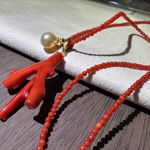 Op de afbeelding: Een koraal ketting met een felrode koraal tak hanger, een parel en een gouden zetting. De ketting is gemaakt van kleine, ronde rode kralen. De sieraden worden gepresenteerd op een houten oppervlak.