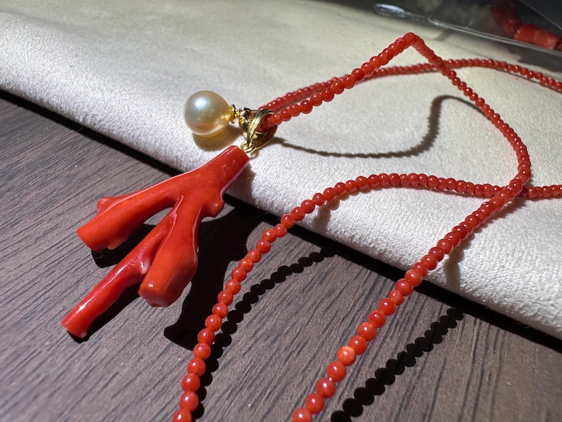 Op de afbeelding: Een koraal ketting met een felrode koraal tak hanger, een parel en een gouden zetting. De ketting is gemaakt van kleine, ronde rode kralen. De sieraden worden gepresenteerd op een houten oppervlak.