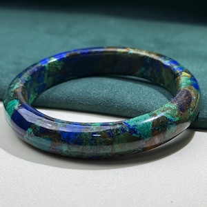 May include: A polished gemstone bangle bracelet. The bracelet features a mosaic of blue, green, and brown hues. The bangle is circular and sits on a white surface with a green backdrop.