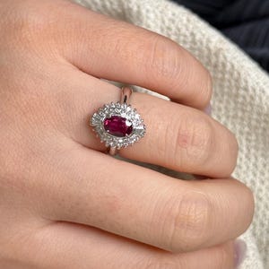 Puede incluir: Un anillo de tono plateado con una piedra preciosa central de color rojo intenso en talla ovalada, rodeada por un halo de pequeñas piedras claras en talla redonda y dos piedras en talla baguette a los lados, sobre una banda de metal pulido.
