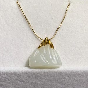 May include: A gold chain necklace with a pendant. The pendant is a white, mountain-shaped stone with gold accents. The necklace is displayed on a white, textured surface.