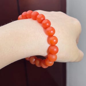May include: A stretch bracelet composed of round, matte-finish beads in a vibrant orange-red hue. The beads are uniform in size and strung together in a continuous circular loop, creating a simple and consistent design around the wrist.