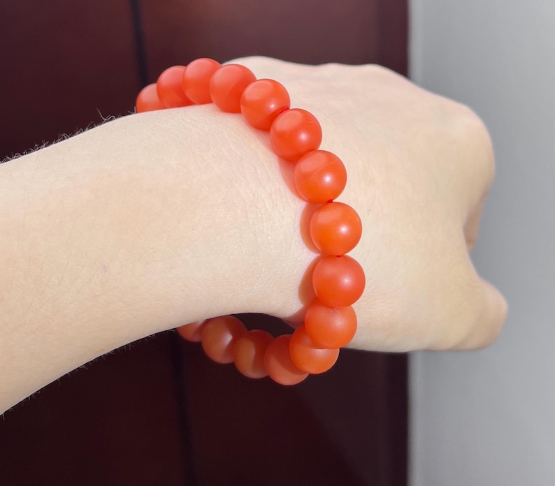May include: A stretch bracelet composed of round, matte-finish beads in a vibrant orange-red hue. The beads are uniform in size and strung together in a continuous circular loop, creating a simple and consistent design around the wrist.