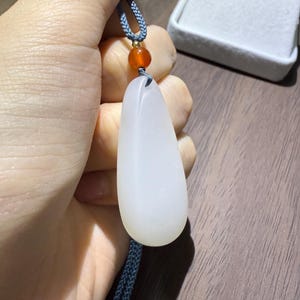 May include: A white jade pendant in a teardrop shape, suspended from a blue cord with a small orange bead. The pendant is smooth and opaque, with a subtle sheen. The cord is a braided design.