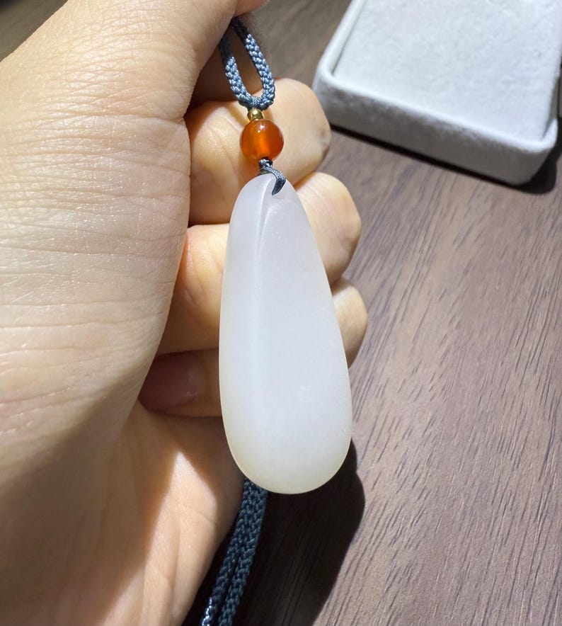May include: A white jade pendant in a teardrop shape, suspended from a blue cord with a small orange bead. The pendant is smooth and opaque, with a subtle sheen. The cord is a braided design.
