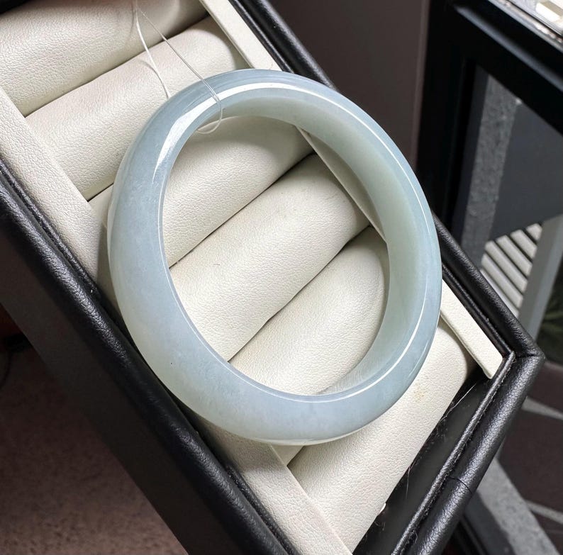 Pode incluir: Um bracelete de jade verde-p&aacute;lido, liso e transl&uacute;cido, com um perfil arredondado. Esta pe&ccedil;a de joalharia circular em pedra apresenta um acabamento polido e varia&ccedil;&otilde;es subtis de cor, exibindo um tom verde-menta suave ao longo da banda.